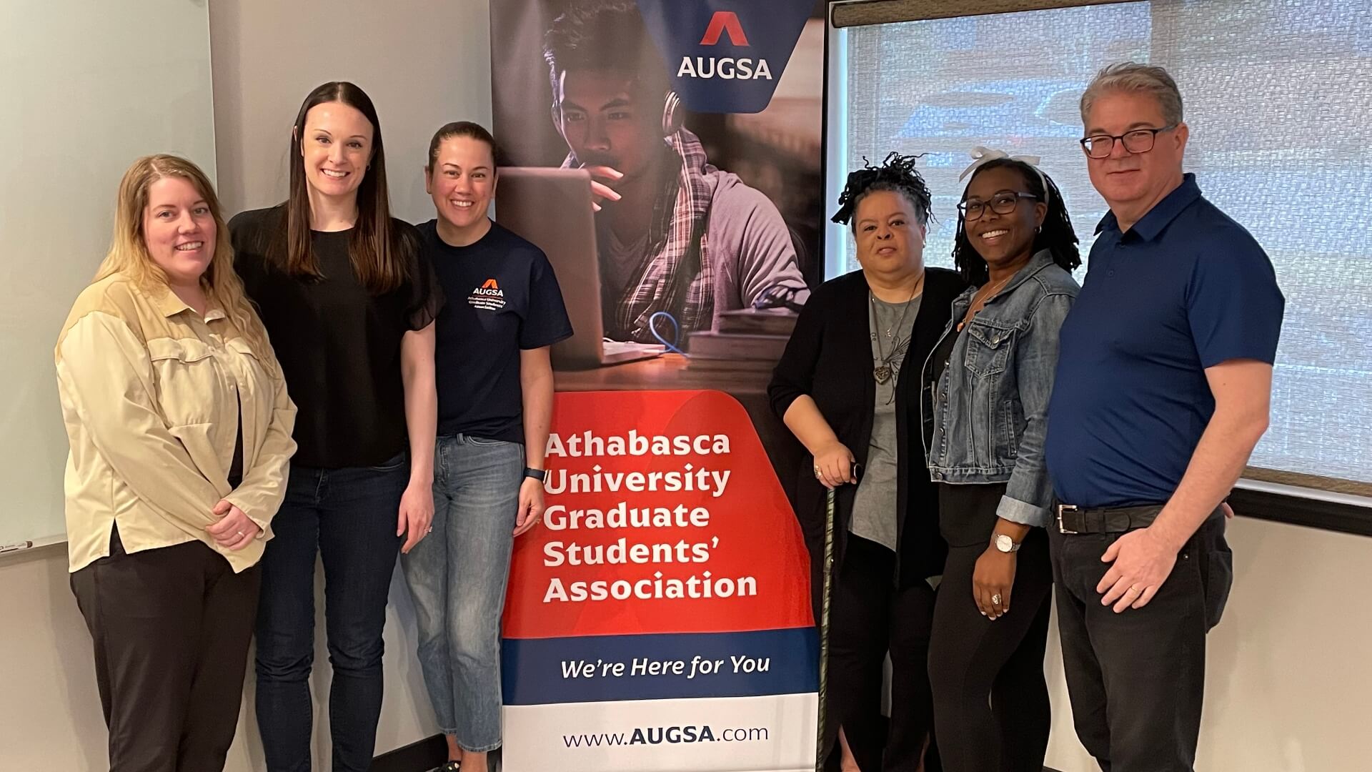 Six members of AUGSA's 24-25 and 25-26 executives standing beside a standup banner promoting AUGSA 