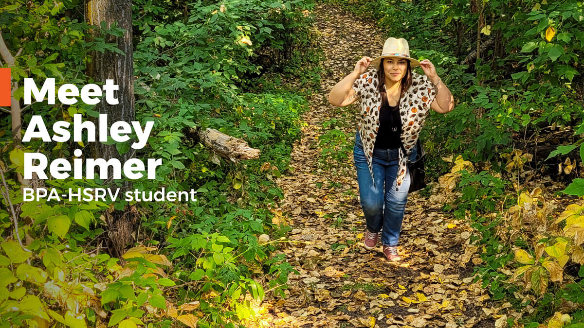Ashley Reimer walking in a forest holding her wide brim hat with both hands